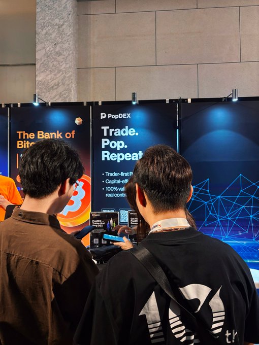 Two people viewed from behind are looking at a PopDEX crypto exchange booth at a conference, with banners reading 'The Bank of Bitcoin' and 'Trade. Pop. Repeat.'