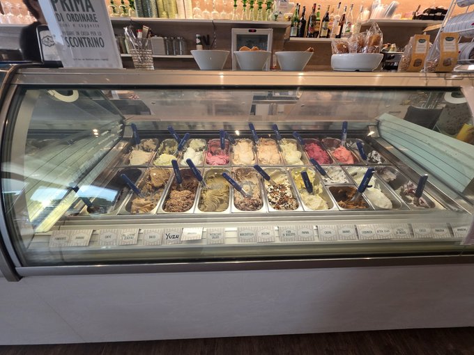 Photograph of a gelato display case filled with various flavors in metal trays, with blue scoops and labeled flavor cards; a sign in the background reads 'PRIMA DI ORDINARE' and 'SCONTRINO'.