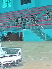 A photo taken from an elevated angle showing a basketball court with several people playing on the upper tier and others sitting on the red stadium seating.