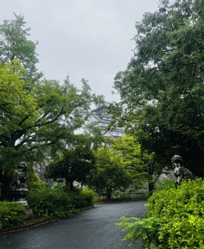 The image shows a park with two statues of lions, one on each side of a path that runs through it. The path appears to be made of asphalt or concrete and is surrounded by lush green trees and bushes. In the background, there are buildings visible behind the foliage, suggesting that this park might be located in an urban area. The post accompanying the image reads "大阪城までayson/stepn", which could possibly be a reference to the name of the park or its location within the city.