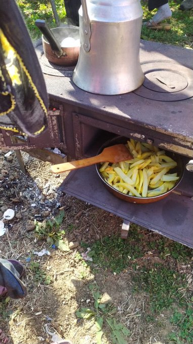 A rustic outdoor cooking scene shows a pan of golden fries with a wooden spoon resting inside, sitting on an old wood stove beside a large metal pot, with someone's foot visible on the ground nearby. The image appears unrelated to the post's discussion of cryptocurrency projects like Axis Robotics, Fhenix, Miden, and BitRobot Network, instead offering a casual, behind-the-scenes glimpse of someone preparing a simple meal outdoors.