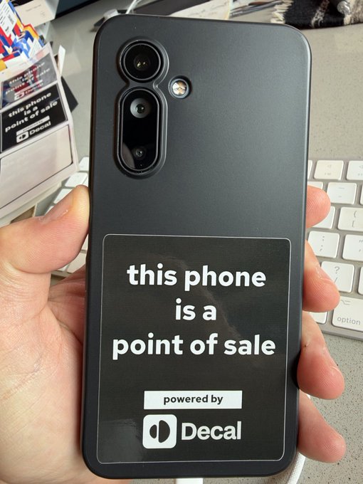A black smartphone with a sticker reading “this phone is a point of sale” and “powered by Decal,” held over papers and a keyboard.
