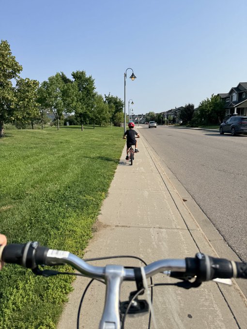 A first-person perspective from a bicycle handlebar following a child in a red helmet riding on a sidewalk along a tree-lined residential street on a sunny day.