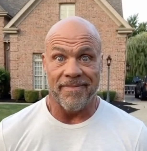 A close-up selfie of a bald man with a gray beard wearing a white t-shirt, making an exaggerated wide-eyed expression in front of a brick house.