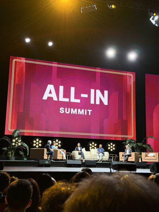 A panel of five speakers sits on stage beneath a large red screen reading "ALL-IN SUMMIT," with an audience watching from the foreground. The post's caption highlights a contrast: the "All-In Summit" features a giant stage screen and a passive, movie-watching audience, suggesting that the upcoming "Bitcoin Alpha" event in October will offer a different, presumably more engaging experience.