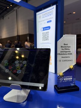 A trade show display for 'CARDANO ACCADEMY' featuring a tablet screen showing transaction icons and an award trophy labeled 'Best Blockchain for RWAs and Agentic Solutions 2026'.