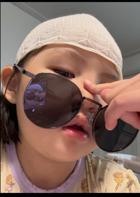 The image features a young girl wearing sunglasses, posing for a picture with her face partially obscured by them. The background of the photo appears to be a room or a living space, as there are chairs present in the scene.