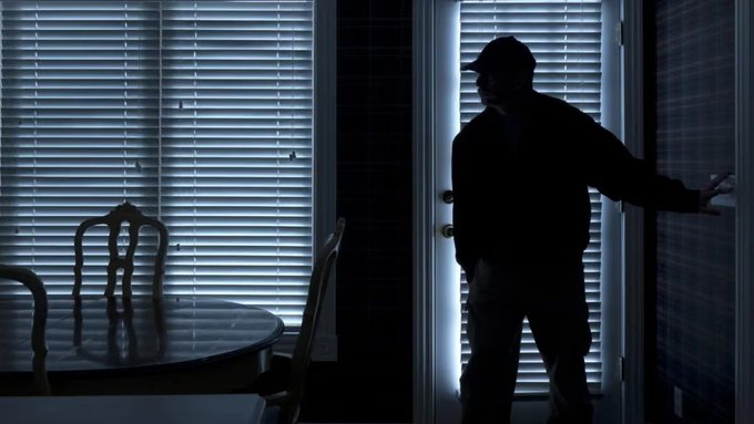 Dark silhouette image of a person wearing a cap standing in a doorway between two rooms with window blinds, next to a table and chair.