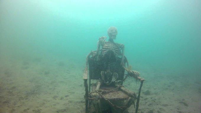 A skeleton sits in a chair underwater, surrounded by murky blue-green water and a sandy seabed.