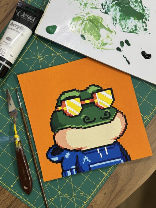 A pixel-art painting of a character with green sunglasses on an orange canvas, sitting on a cutting mat with acrylic paint and brushes nearby.