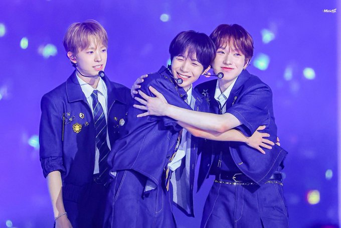Three performers in dark blue uniforms embrace warmly on stage during a concert with a purple, bokeh-lit background.