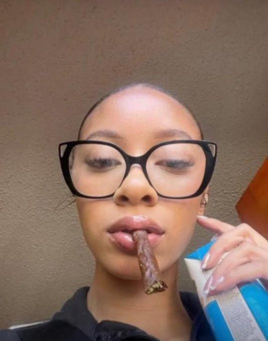 A close-up selfie of a young woman with glasses smoking a cigar while holding a blue and white bag, appearing to film for social media.
