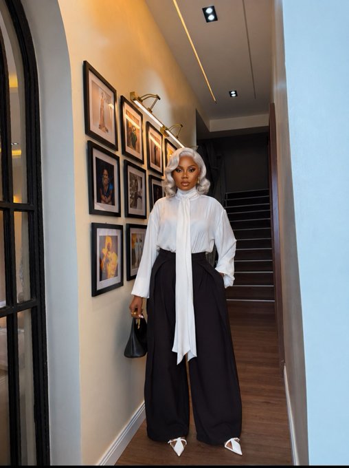 A woman with silver-white hair wears a white blouse with a long tie and black wide-leg pants, posing in a hallway lined with framed photographs.