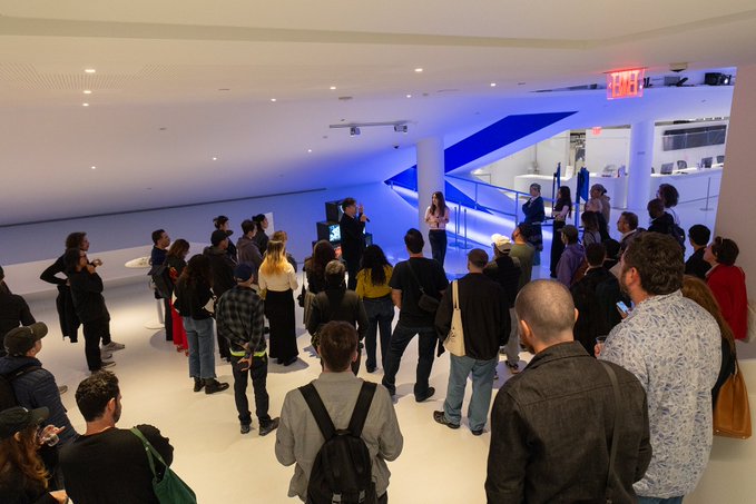 A crowd of people gathered in a modern, white-walled retail space with blue accent lighting, listening to a presentation or tour.