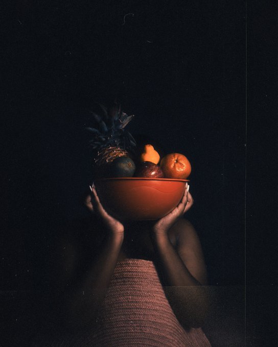 A dark, moody photograph of a person holding an orange bowl filled with fruit (including a pineapple and oranges) above their head, obscuring their face.