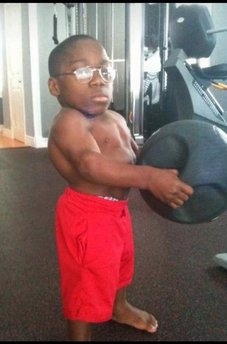 A young boy wearing glasses and red shorts holds a large black medicine ball in a gym setting.
