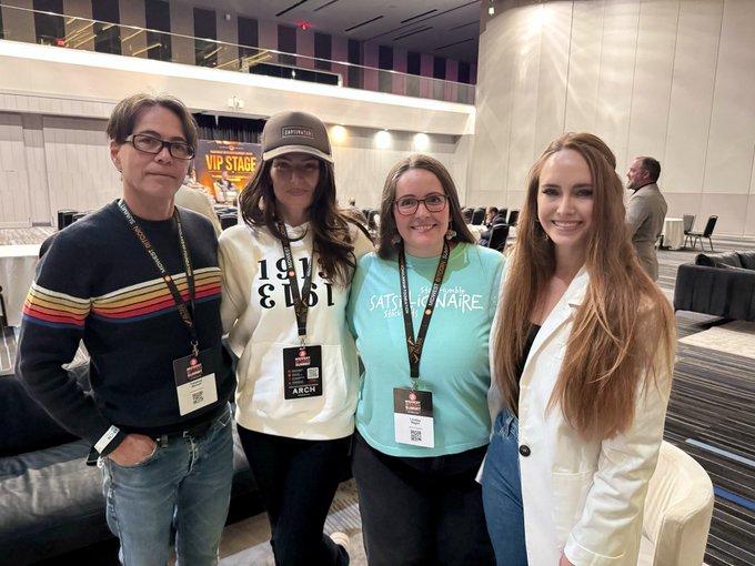 Four people pose together at what appears to be a conference or event venue, with some wearing lanyards with 'ARCH' badges and one wearing a 'SATELLIONAIRE' t-shirt.