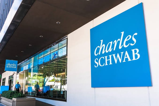 A Charles Schwab branch exterior features a large blue sign with white 'charles SCHWAB' text alongside a smaller matching sign near the entrance, with glass windows reflecting the surroundings.