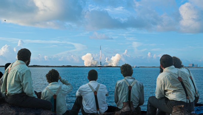 A group of people in white shirts and suspenders sitting on a shoreline watching a SpaceX rocket launch in the distance across the water.