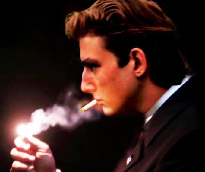 A young man in a dark suit is shown in profile, lighting a cigarette with a lighter against a dark background.