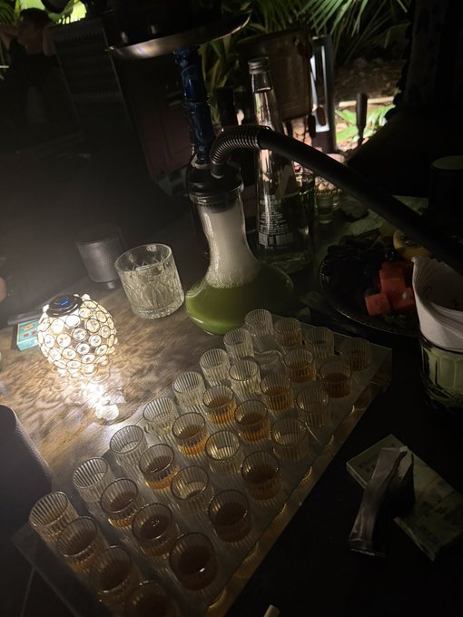 A dimly lit bar setup with a tray of shot glasses containing amber liquid, a glass with a green beverage, bottles, and a decorative glowing lamp on a wooden counter.