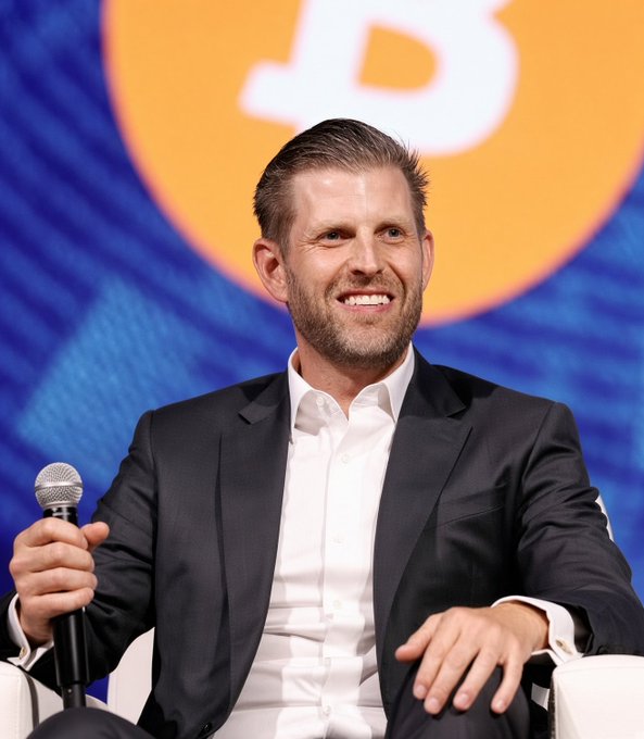 A smiling man in a dark suit and white shirt holds a microphone while seated on stage at what appears to be a cryptocurrency conference, with a partial Bitcoin logo visible in the background. The image relates to the post as it shows Eric Trump, co-founder of the Trump family-backed crypto venture WLFI, discussing the company's interest in using stablecoins to pay employees.