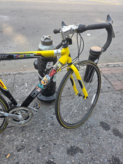 A yellow Trek road bike with stickers parked and locked to a black fire hydrant on a city street.