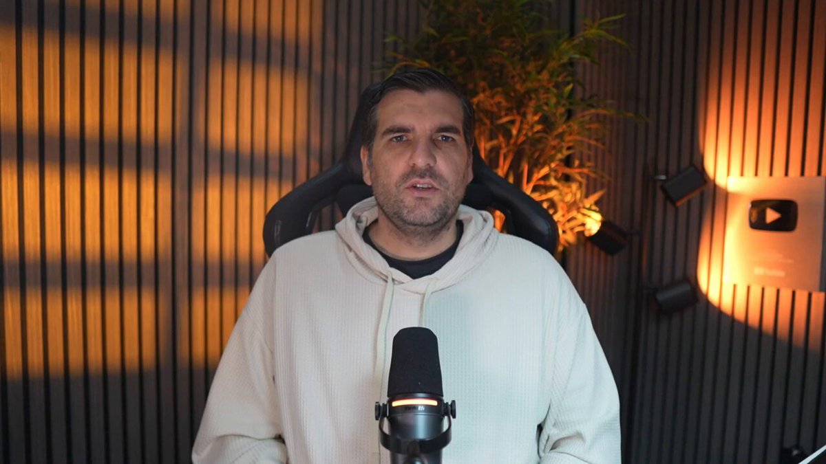 The image shows a man wearing a white hoodie sitting behind a microphone with his hands on his knees. He appears to be giving a speech or presentation, possibly at an event like a podcast or webinar. The background features a wall with a window that allows natural light into the room and casts shadows onto the man's face. There is also a potted plant in the scene, adding some greenery to the environment.