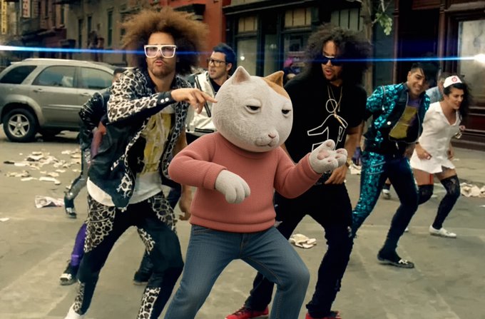 An LMFAO-style music video scene features a person wearing a giant cartoon cat head and a pink sweater dancing on a city street alongside several backup dancers in eccentric outfits.