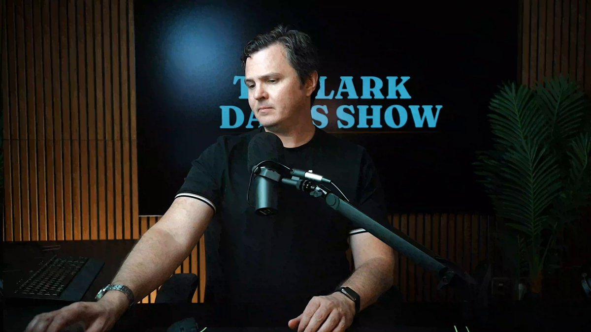 The image features a man sitting at a desk with a microphone, likely preparing for a podcast or radio show. The background includes a TV screen displaying "The Clark's Show" and a plant nearby. There are also some text elements on the image that read "URGENT! Bitcoin In The Buy Zone 0:00 Bitcoin Buy Zone 1:42 Bitcoin Bottom 6:22 Ethereum Updates 6:58 Crypto Rug 7:58 ICO Facts".