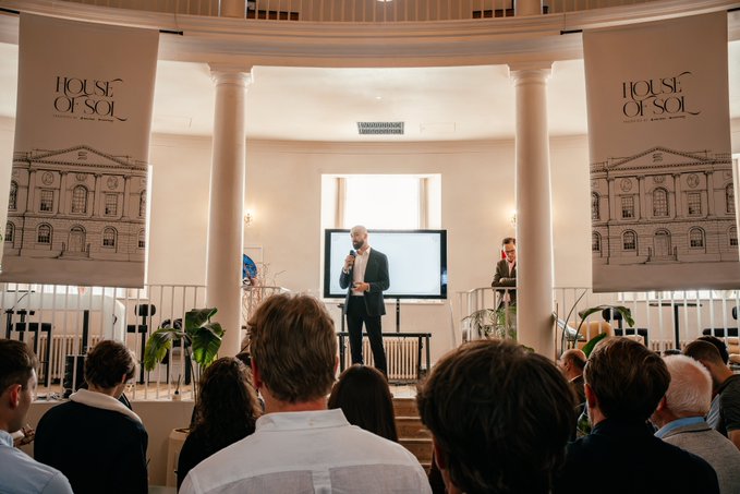 An indoor event in a circular room with columns where a man in a suit presents on stage, flanked by 'HOUSE OF SOL' banners, to an audience seated in the foreground.