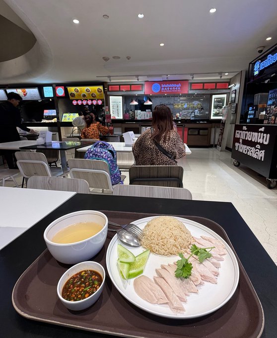 A $2.86 Hainanese chicken rice meal from KFC sits on a tray in what appears to be a Bangkok food court, paired with broth and chili sauce. The scene captures a casual mall dining setting where the poster is enjoying affordable food while coding onchain apps, as reflected in the background food stalls and signage.