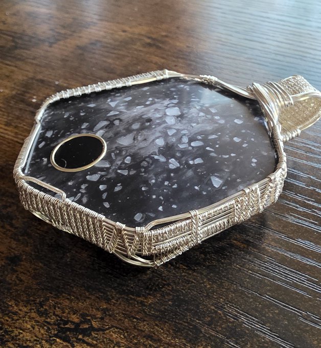 A polished black stone pendant with white speckles, accented by a small black onyx cabochon and intricate silver wire wrapping, rests on a wooden surface. The piece embodies self-custody as a statement, pairing the rugged beauty of natural stone with meticulous craftsmanship to symbolize independence and the secure ownership of a Bitkey wallet.