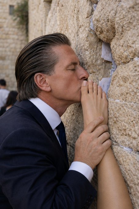 A man in a suit pressing his face and hands against the Western Wall in Jerusalem, kissing a note tucked between the ancient stones in prayer.