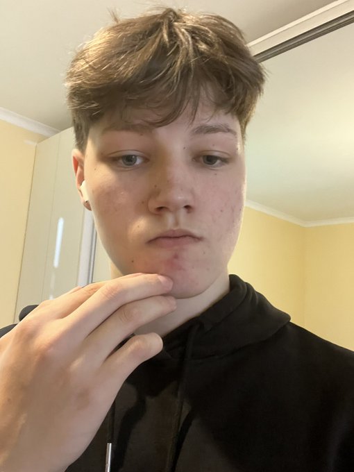 A young person with short brown hair wearing a black hoodie, posing thoughtfully with their hand near their chin.