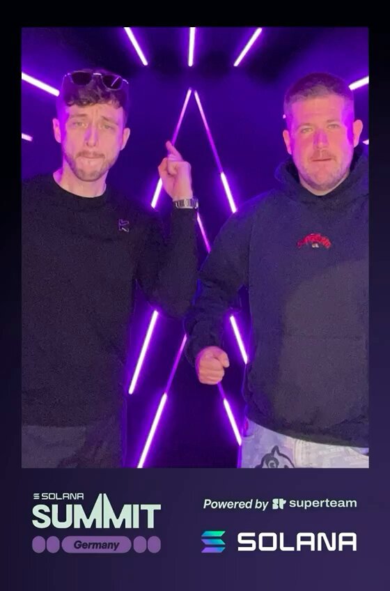 The image features two men standing next to each other, both wearing black shirts with purple designs. They are positioned against a backdrop of purple lights that create an interesting visual effect. The text "Bosh from Solana Summit" can be seen on the image, which suggests that this photo was taken at the Solana Summit event.