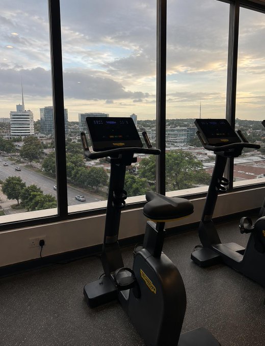 Two Technogym stationary exercise bikes face floor-to-ceiling windows overlooking a city skyline at dusk.