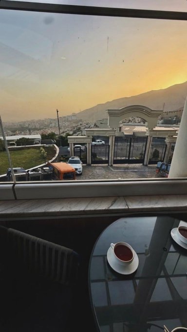 A cozy morning view through a window shows a small table with two cups of tea in the foreground, overlooking a gated entrance and distant cityscape bathed in soft sunrise light—capturing a peaceful "good morning" moment to start the new week.