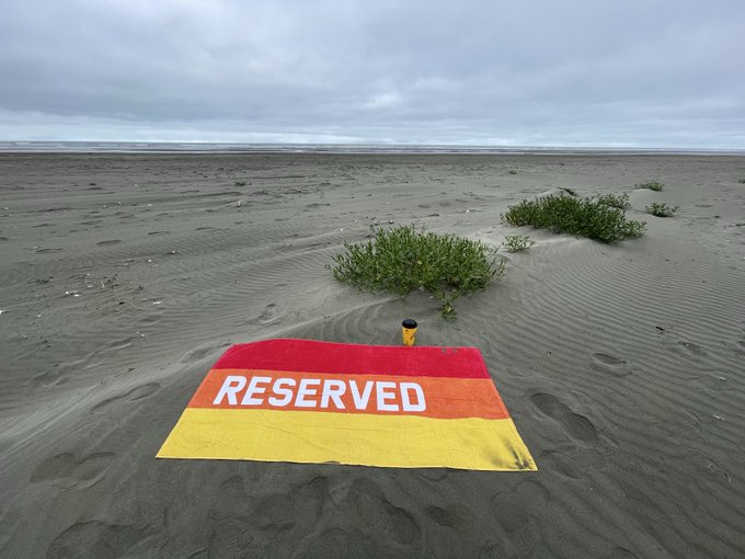 A towel marked 'RESERVED' with orange, yellow, and red stripes lies on a sandy beach under a cloudy sky, near some small green plants.