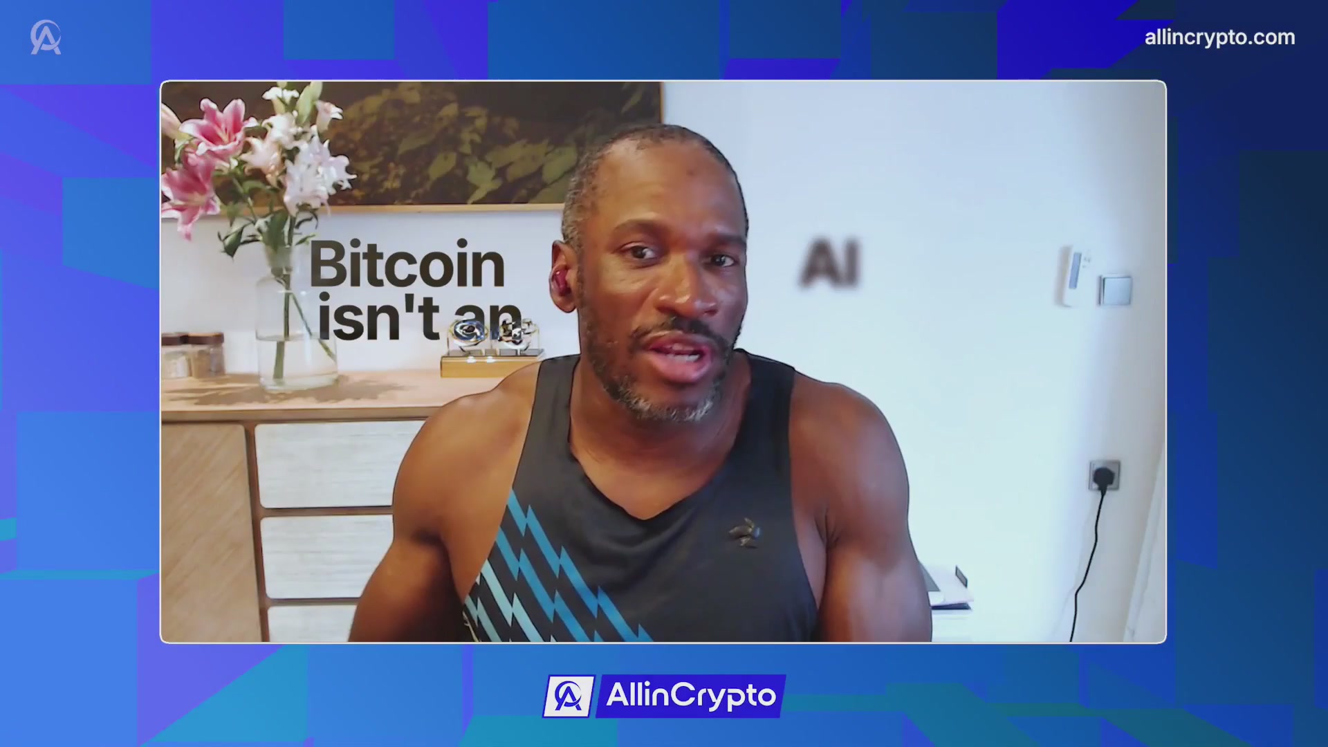The image shows a man wearing a black tank top with blue stripes sitting at a desk while talking into a microphone during an online meeting or webinar. The background of the image features a blue wall, which has a poster that says "BITCOIN ISN'T AN AI". There is also a vase with flowers on the desk next to the man.