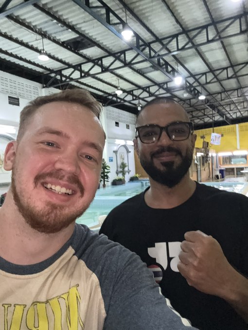 Two men pose for a selfie at what appears to be an indoor swimming pool venue, one wearing glasses and a black t-shirt with a fist raised, the other in a grey and white shirt smiling at the camera.