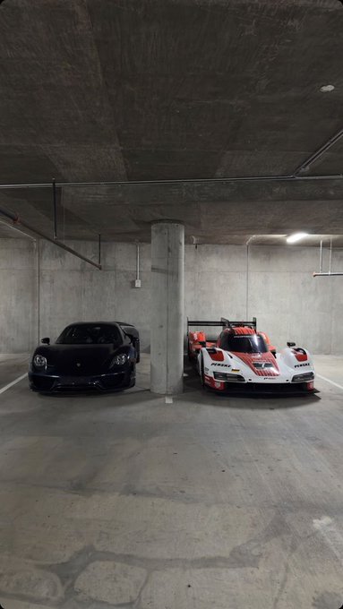 Two sports cars parked in a concrete parking garage: a black Porsche on the left and a white, red, and black Porsche race car (Porsche motorsport livery) on the right.