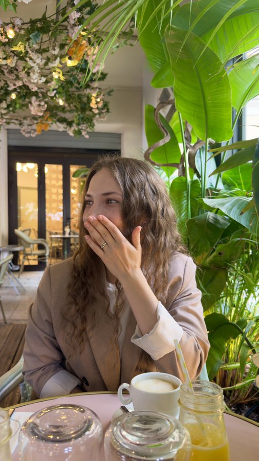 A woman with curly hair in a beige blazer sits at a cafe table, covering her mouth with her hand, with a coffee cup and drinks in the foreground and plants in the background.