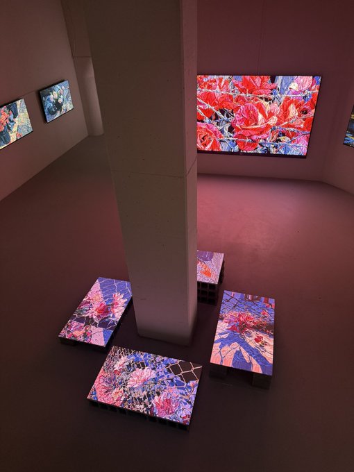 An art gallery installation displays vibrant red and blue floral patterned artworks on wall-mounted screens and low platforms around a central white column.