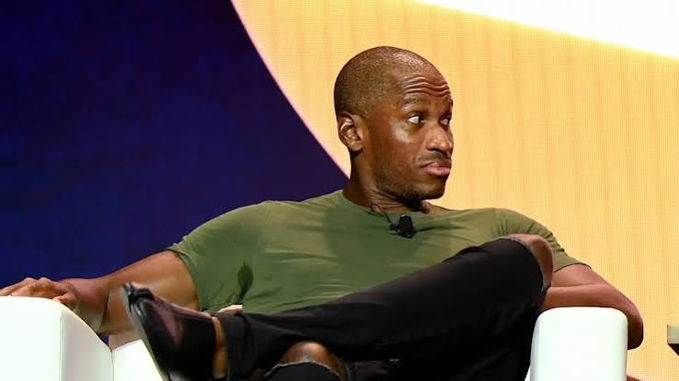A bald Black man in a green t-shirt and black pants sits casually in a white chair against a blue and yellow backdrop.