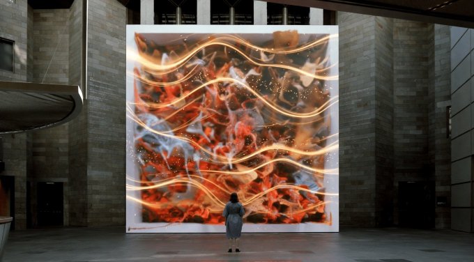 A person stands in a large concrete atrium viewing a massive, glowing digital art installation displaying swirling orange and white flame-like patterns.