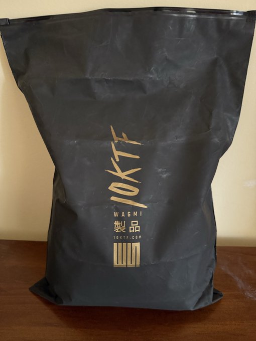 A black packaging bag with gold '10KTF WAGMI' branding and Japanese characters (製品) sitting on a wooden surface against a beige wall.