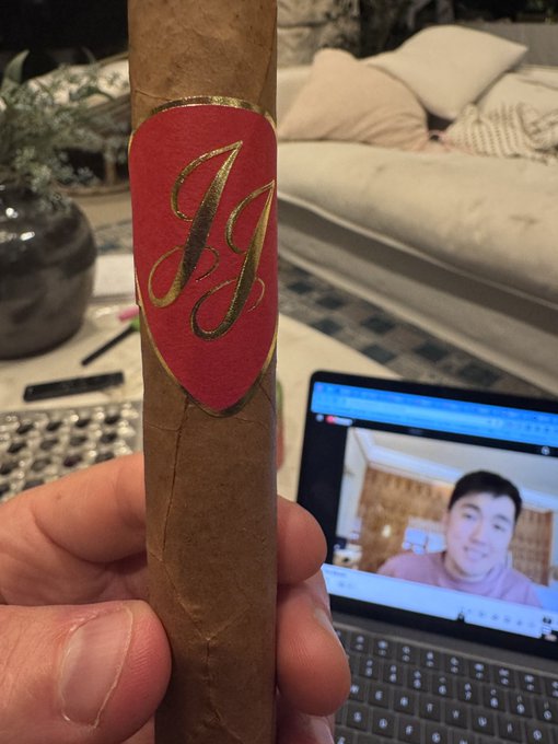 A close-up of a hand holding a cigar with a red and gold band featuring a stylized 'H' monogram, with a laptop showing a video call in the background.