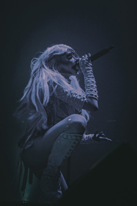 A dramatic concert photograph showing a female performer with long blonde hair singing into a microphone under blue stage lighting.