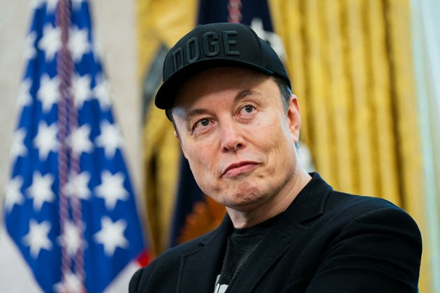 Elon Musk, wearing a black "DOGE" cap and blazer, is pictured in front of American and presidential flags, visually grounding his bold forecast that AI and robots will more than double the global economy within the decade. His expression and the official backdrop lend weight to the claim that AI is poised to drive one of history's largest economic transformations.
